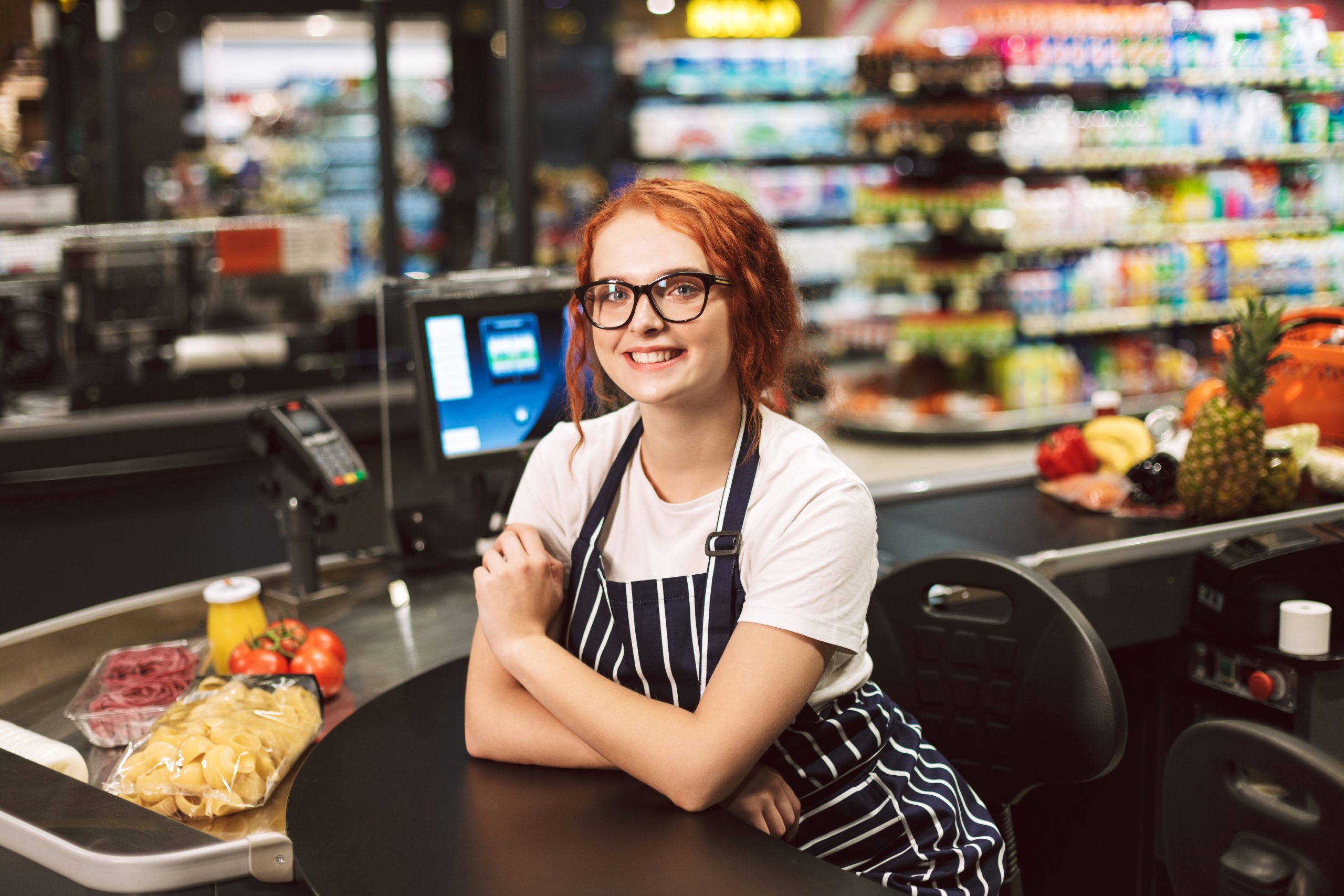 Pretty smiling cashier in eyeglasses and striped apron happily looking in camera working in modern supermarket